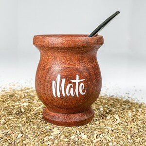 May include: A brown wooden mate gourd with the word "Mate" written in white on the side. The gourd is sitting on a bed of dried yerba mate leaves.