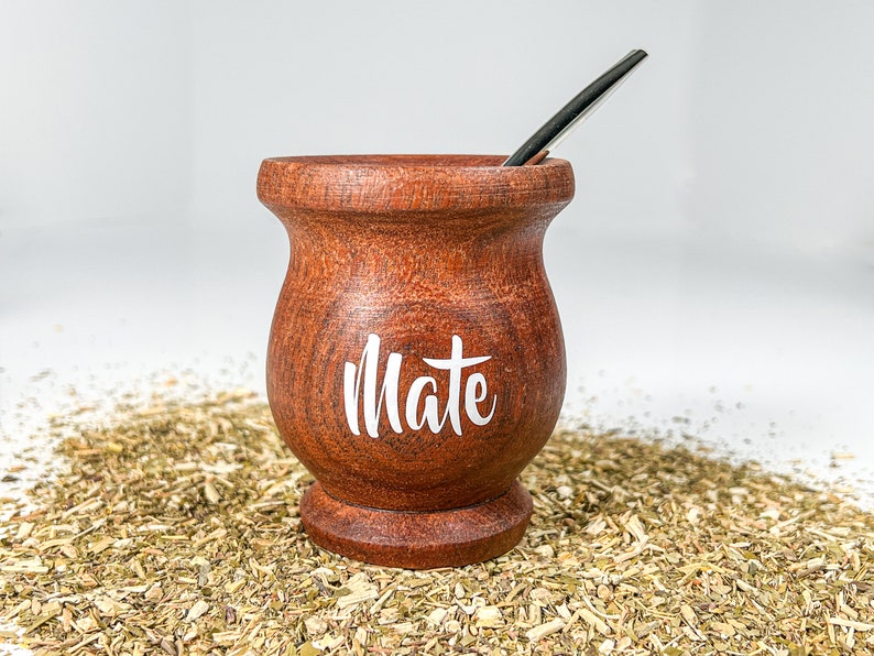 May include: A brown wooden mate gourd with the word "Mate" written in white on the side. The gourd is sitting on a bed of dried yerba mate leaves.