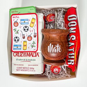 May include: A white gift box filled with a wooden mate cup, a silver bombilla straw, a red and white candy bar, a small round tin, and a package of mate tea. The mate tea package has a red border and the text "CRUZ DE MALTA El sabor de la tradici&oacute;n" in Spanish. The package also has the text "CONT NETO 500g NET WEIGHT 17.6 Oz" in English and Spanish.
