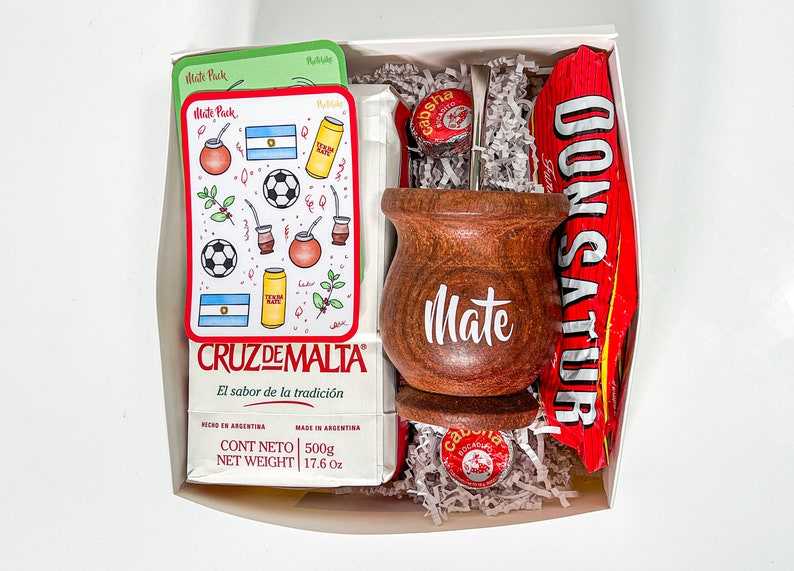 May include: A white gift box filled with a wooden mate cup, a silver bombilla straw, a red and white candy bar, a small round tin, and a package of mate tea. The mate tea package has a red border and the text "CRUZ DE MALTA El sabor de la tradici&oacute;n" in Spanish. The package also has the text "CONT NETO 500g NET WEIGHT 17.6 Oz" in English and Spanish.