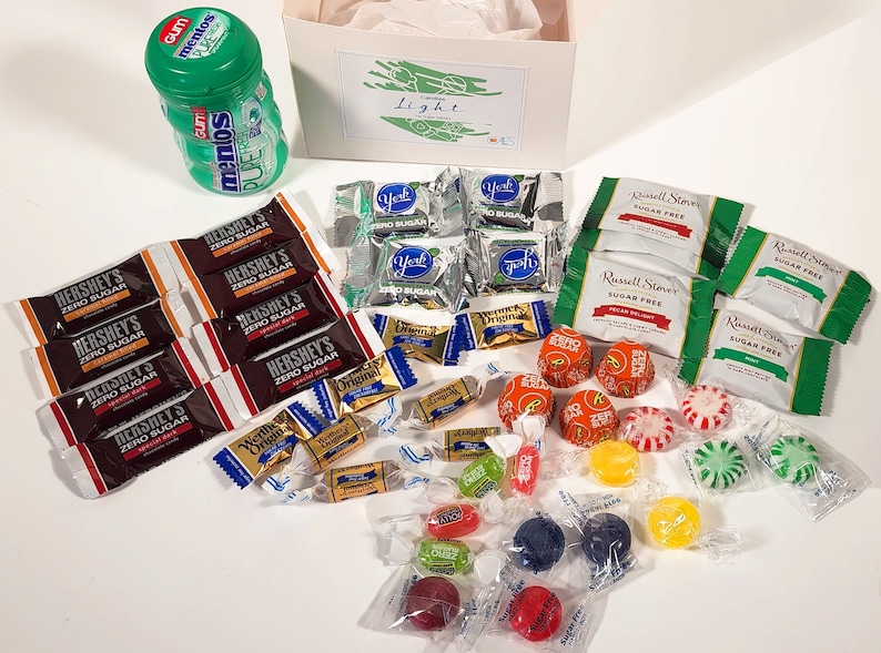 May include: A white gift box with a green and white label that says "Light" on it. The box is filled with a variety of candies, including chocolate bars, hard candies, and gum. The candies are wrapped in colorful wrappers.