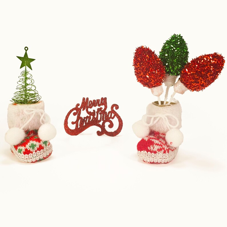 May include: Two red and white knitted boot-shaped Christmas decorations. One decoration contains a small green Christmas tree and the other has three red and green fluffy baubles inside. The words "Merry Christmas" are written in red glitter on a white background.