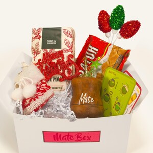 May include: A white gift box with a red and white "Mate Box" logo. The box contains a variety of items, including a red and green Christmas ornament, a brown wooden mate cup, a bag of mate tea, a red and white bag of sugar, and a green and white card with instructions for making mate.