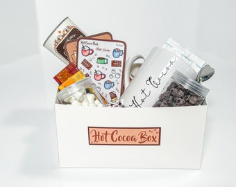 May include: A white gift box with the text "Hot Cocoa Box" on the front. The box is filled with a white mug that says "Hot Cocoa", a jar of marshmallows, a bag of chocolate chips, a bag of hot cocoa mix, and a small spoon.