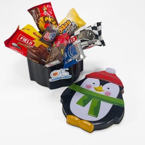 May include: A black container filled with various snack packages including Field Choko Soda, Picaras, and Sublime, next to a penguin-shaped metal lid featuring a red hat, green scarf, and yellow beak on a white background.