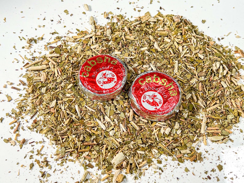 May include: Two red and silver foil-wrapped packages of yerba mate tea, labelled "Caba&ntilde;a", on a bed of loose dried yerba mate leaves.