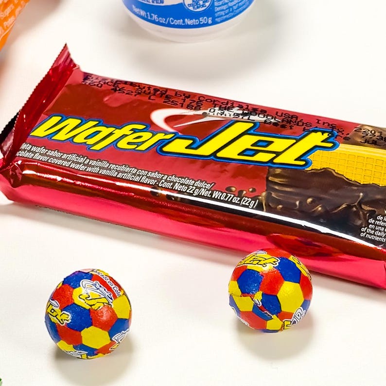 May include: A red and yellow "Wafer Jet" candy bar with a chocolate and vanilla flavor, alongside two small, colorful soccer ball-shaped candies. The candy bar is in a red wrapper with text. The candies are on a white surface.