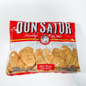 May include: A red and white bag of Don Satur biscuits, a brand of savoury crackers. The bag features the brand name "Don Satur" in large red letters, the words "Fundado en 1967" in smaller red letters, and a picture of a man with a moustache. The bag contains 200 grams of biscuits and the text "Bizcocho Salado" is printed on the front of the bag.