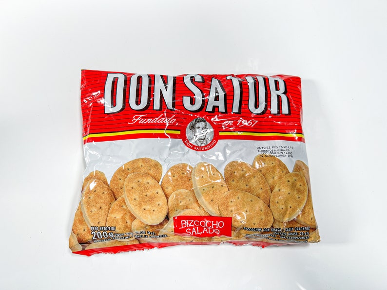 May include: A red and white bag of Don Satur biscuits, a brand of savoury crackers. The bag features the brand name "Don Satur" in large red letters, the words "Fundado en 1967" in smaller red letters, and a picture of a man with a moustache. The bag contains 200 grams of biscuits and the text "Bizcocho Salado" is printed on the front of the bag.