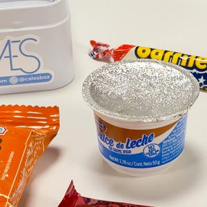 May include: A close-up of a small container of "Dulce de Leche" caramel spread, with a foil lid. The container is white with blue and brown accents. A wrapped candy bar and a partially visible orange package are also in the image.