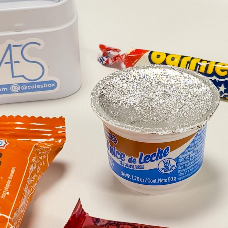 May include: A close-up of a small container of "Dulce de Leche" caramel spread, with a foil lid. The container is white with blue and brown accents. A wrapped candy bar and a partially visible orange package are also in the image.