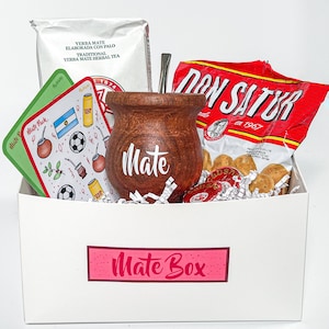 Peut inclure: Une boîte cadeau blanche avec une étiquette rose qui dit "Mate Box". La boîte contient une gourde à mate en bois brun, une bombilla argentée, un sac de yerba mate, un sac de biscuits et une carte avec un ballon de football et le drapeau argentin.