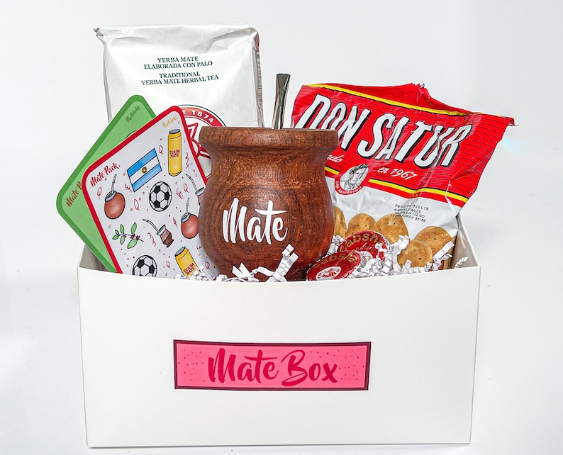May include: A white gift box with a pink label that says "Mate Box". The box contains a brown wooden mate gourd, a silver bombilla, a bag of yerba mate, a bag of biscuits, and a card with a football and the Argentine flag.