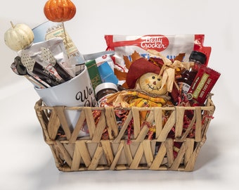 Può includere: Un cesto intrecciato pieno di articoli a tema autunnale. Include uno spaventapasseri, decorazioni a forma di zucca, una tazza con la scritta "Warm Up", snack e sciroppo d'acero. Il cesto è riempito con carta sminuzzata rossa e arancione.