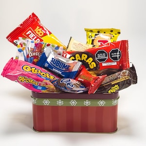 May include: A red and maroon tin filled with various wrapped candies. The candies are in colorful wrappers with text such as "FIELD", "Glacitas", and "SUBLIME". The tin has a green ribbon with white snowflake designs.