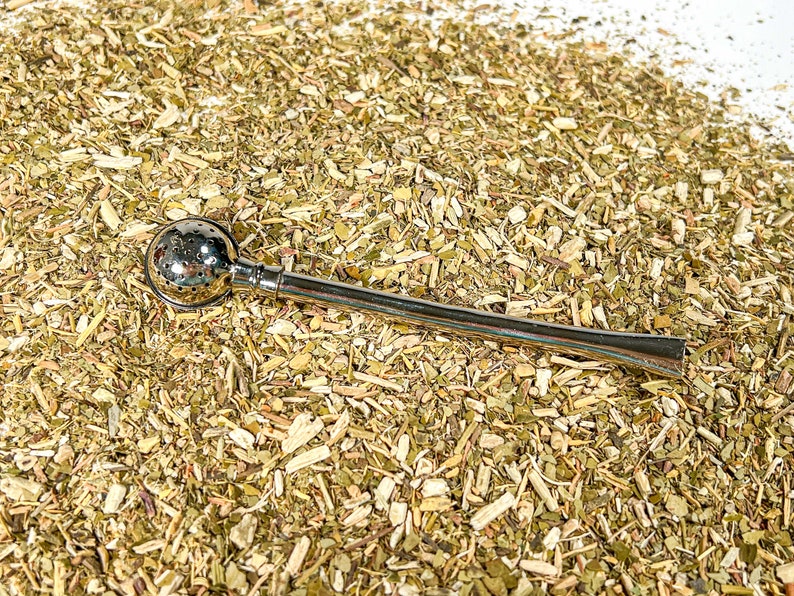 May include: A silver bombilla, a traditional tool for drinking yerba mate, sits on a bed of dried yerba mate leaves.
