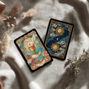 Puede incluir: Dos cartas de tarot con dise&ntilde;os ornamentados. Una carta presenta una paloma, un c&aacute;liz dorado y rosas. La otra representa dos soles, nubes y una luna creciente. Ambas cartas tienen bordes negros y detalles intrincados.