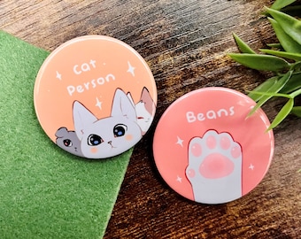 Könnte beinhalten: Zwei runde rosa und orangefarbene Knöpfe mit weißen Bildern. Der erste Knopf zeigt eine weiße Katze mit dem Text "Cat Person" in Schwarz. Der zweite Knopf zeigt eine weiße Pfote mit dem Text "Beans" in Schwarz.
