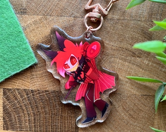 Könnte beinhalten: Ein durchsichtiger Acryl-Schlüsselanhänger mit einem goldenen sternförmigen Verschluss, auf dem ein roter und schwarzer Comic-Charakter mit Hörnern und einem Mikrofon abgebildet ist.