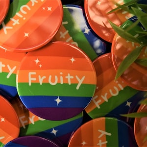 Op de afbeelding: Een close-up van ronde pinnen in regenboogkleuren met het woord "Fruity" in wit op elke pin. De pinnen zijn verspreid over een oppervlak met groene bladeren.