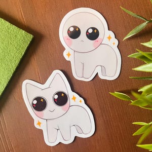 Peut inclure: Deux autocollants de dessin animé blancs avec de grands yeux noirs et des détails scintillants. Un autocollant est un personnage rond avec une expression surprise. L'autre autocollant est un personnage ressemblant à un chat avec une expression joyeuse.