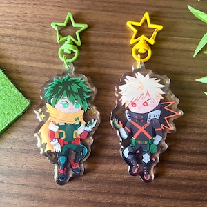 Op de afbeelding: Twee acryl sleutelhangers met anime-personages. Eén sleutelhanger heeft een groenharig personage in een groene, gele en rode outfit. De andere sleutelhanger heeft een stekelig blond personage in een zwart-oranje outfit. Beide hebben stervormige sluitingen.