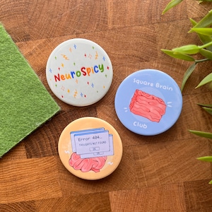 May include: Three round pins with colorful designs. The first pin is white with a rainbow colored text that says "NeuroSpicy" and colorful stars. The second pin is blue with a pink brain shaped like a square and the text "Square Brain Club". The third pin is yellow with a pink brain and a blue computer screen with the text "Error 404... Thoughts not found".