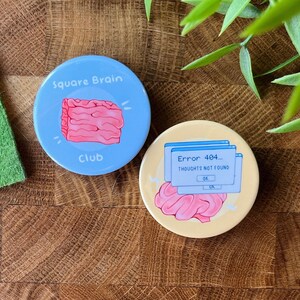 May include: Two round buttons with a light blue and yellow background. The blue button has a pink square brain with the text "Square Brain Club". The yellow button has a pink brain with a blue computer error message that says "Error 404... Thoughts not found".