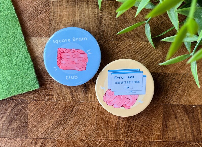 May include: Two round buttons with a light blue and yellow background. The blue button has a pink square brain with the text "Square Brain Club". The yellow button has a pink brain with a blue computer error message that says "Error 404... Thoughts not found".