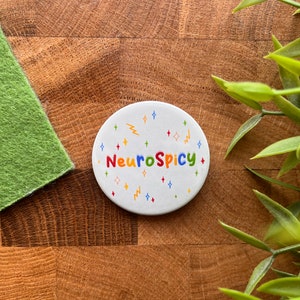 May include: White round sticker with the text "NeuroSpicy" in colorful letters. The sticker is decorated with colorful stars and lightning bolts.
