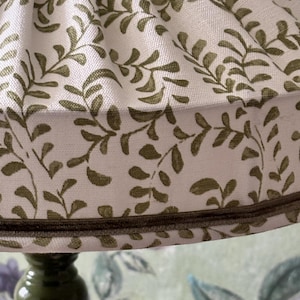 May include: A close-up of a lampshade with a cream-colored background and a repeating pattern of olive-green leaves and vines. The lampshade is trimmed with a dark brown border and sits atop a dark green lamp base.