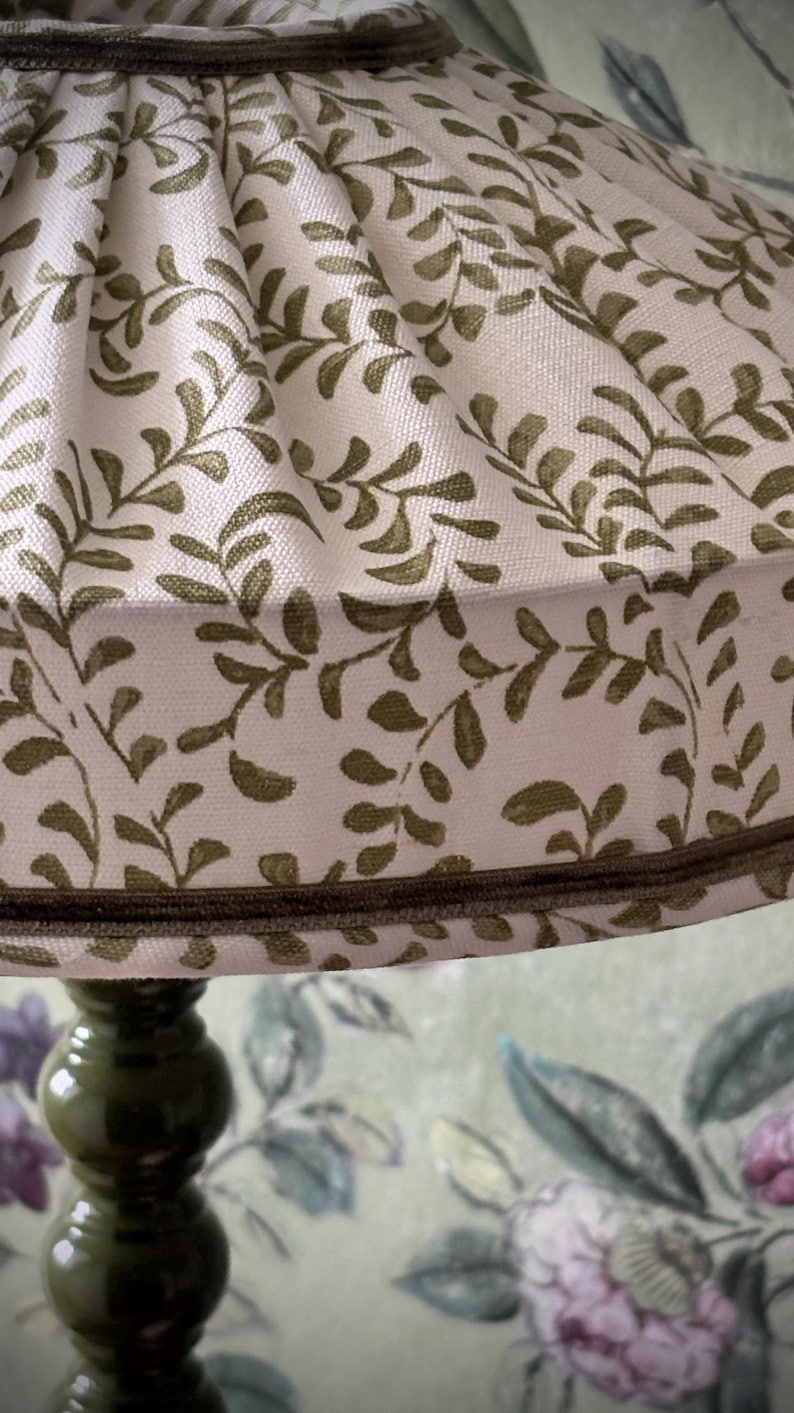 May include: A close-up of a lampshade with a cream-colored background and a repeating pattern of olive-green leaves and vines. The lampshade is trimmed with a dark brown border and sits atop a dark green lamp base.