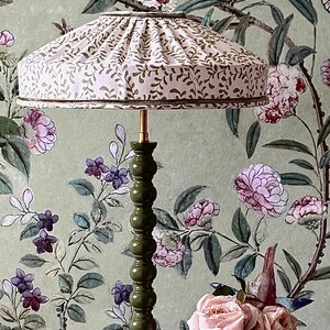 May include: A decorative table lamp with a green base and a cream-colored lampshade adorned with a brown leaf pattern. The lamp is set against a floral wallpaper background, with a bouquet of pink roses on a wooden surface.
