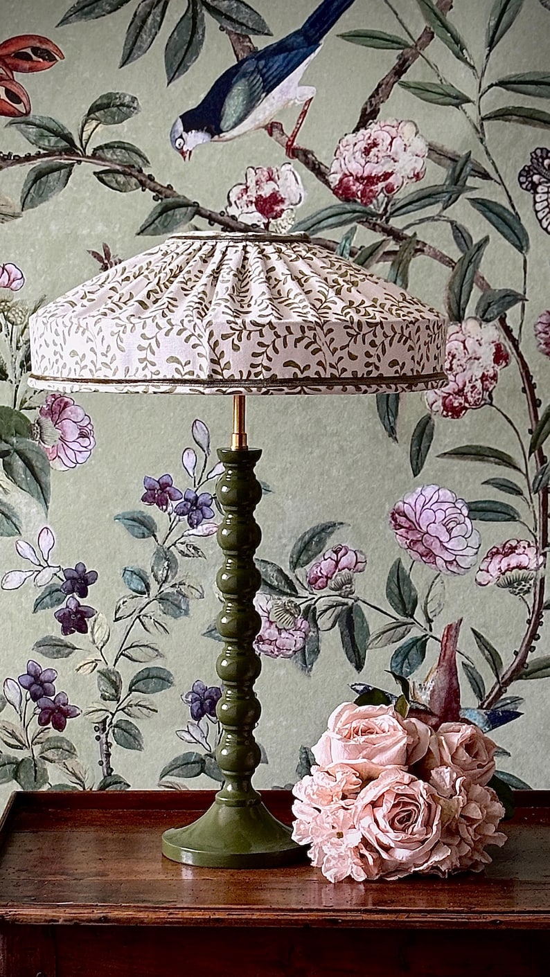 May include: A decorative table lamp with a green base and a cream-colored lampshade adorned with a brown leaf pattern. The lamp is set against a floral wallpaper background, with a bouquet of pink roses on a wooden surface.