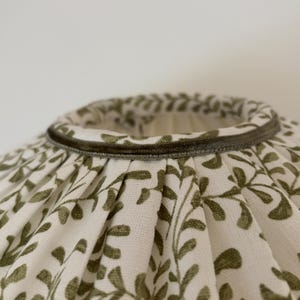 May include: A lampshade with a white background and a repeating pattern of olive green leaves. The top edge is trimmed with a dark green velvet-like material.