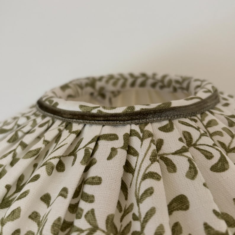 May include: A lampshade with a white background and a repeating pattern of olive green leaves. The top edge is trimmed with a dark green velvet-like material.