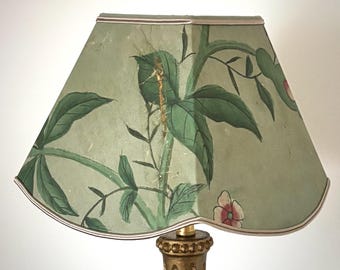 May include: A lampshade with a light green background and a botanical design featuring green leaves and pink flowers. The lampshade has a scalloped edge and a gold-colored trim. The lamp base is gold-colored.