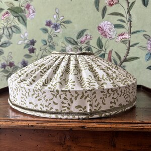 May include: A cream-colored lampshade with a green leaf pattern. The lampshade has a rounded shape and is trimmed with a green border. The background features a floral wallpaper.