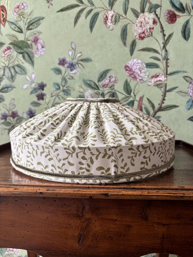 May include: A cream-colored lampshade with a green leaf pattern. The lampshade has a rounded shape and is trimmed with a green border. The background features a floral wallpaper.