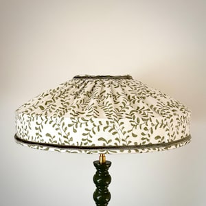 May include: A decorative lampshade with a white background and a repeating pattern of olive green leaves. The lampshade has a dark green trim and sits atop a dark green lamp base.