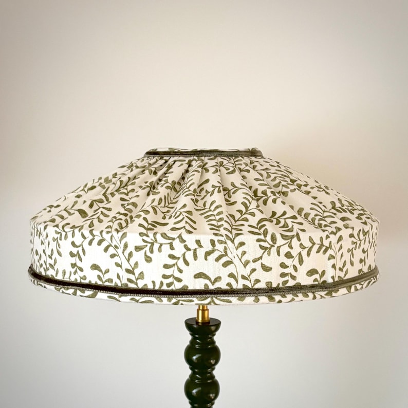 May include: A decorative lampshade with a white background and a repeating pattern of olive green leaves. The lampshade has a dark green trim and sits atop a dark green lamp base.