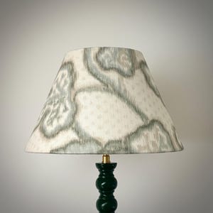 May include: A lampshade with a cream-colored background and a pattern of gray and light blue shapes. The lampshade sits atop a dark green lamp base with a brass fitting.