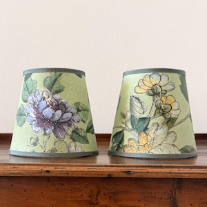 Peut inclure: Deux petits abat-jours coniques avec des motifs floraux. Les abat-jours ont un fond vert clair avec des illustrations de fleurs violettes et jaunes et de feuilles vertes. Les bords sont bordés d'un ruban gris-vert. Ils sont posés sur une surface en bois.