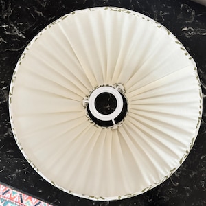 May include: Cream-colored lampshade with a pleated design. The shade has a white metal frame and a decorative border with green leaf patterns. The lampshade is round and appears to be made of fabric.