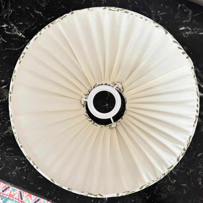 May include: Cream-colored lampshade with a pleated design. The shade has a white metal frame and a decorative border with green leaf patterns. The lampshade is round and appears to be made of fabric.