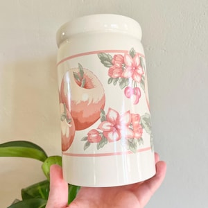 May include: Cream-colored ceramic jar with a lid, featuring a decorative band with peach-colored apples, pink flowers, and cherries. The jar is held in a hand, showcasing its cylindrical shape and floral design. A vintage-style kitchenware item.