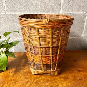 May include: A tall, woven wicker basket with a rectangular base and a flared top. The basket features vertical and horizontal woven patterns in shades of brown, orange, and tan. The basket is sitting on a wooden surface.