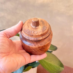 May include: A small, round wooden container with a lid, held in a hand. The container is brown with a textured surface and the word "MAYONNAISE" is carved into the side. The lid has a small, cylindrical handle.