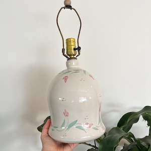 May include: A bell-shaped ceramic lamp base with a glossy off-white finish, featuring a hand-painted pattern of small pink tulip-like flowers and green leaves. The lamp includes a brass-colored metal socket and a wire harp with a wooden finial.