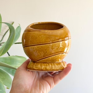 May include: A small, round, ceramic vase in a warm, mustard yellow hue. The vase features a series of diagonal, raised lines encircling its body. It sits on a decorative, pedestal base. The opening is oval-shaped.